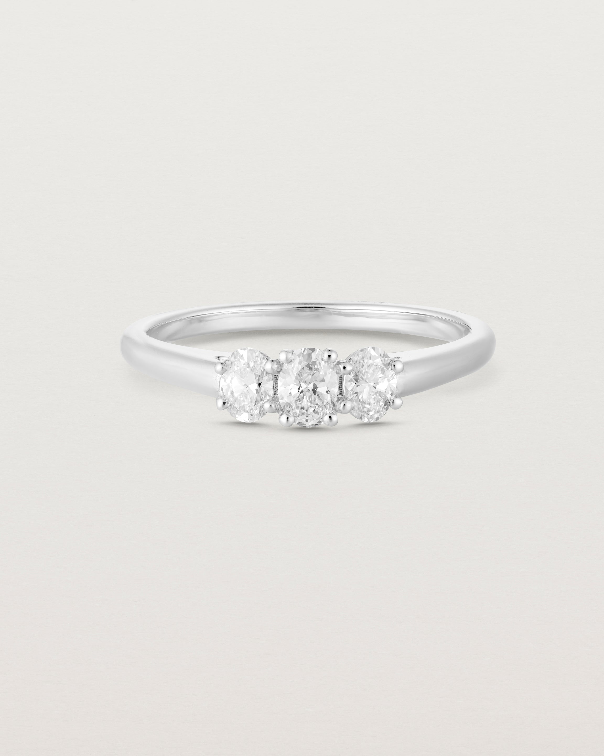 Front on deep etched image of a white gold engagement ring with three oval white laboratory grown diamonds.