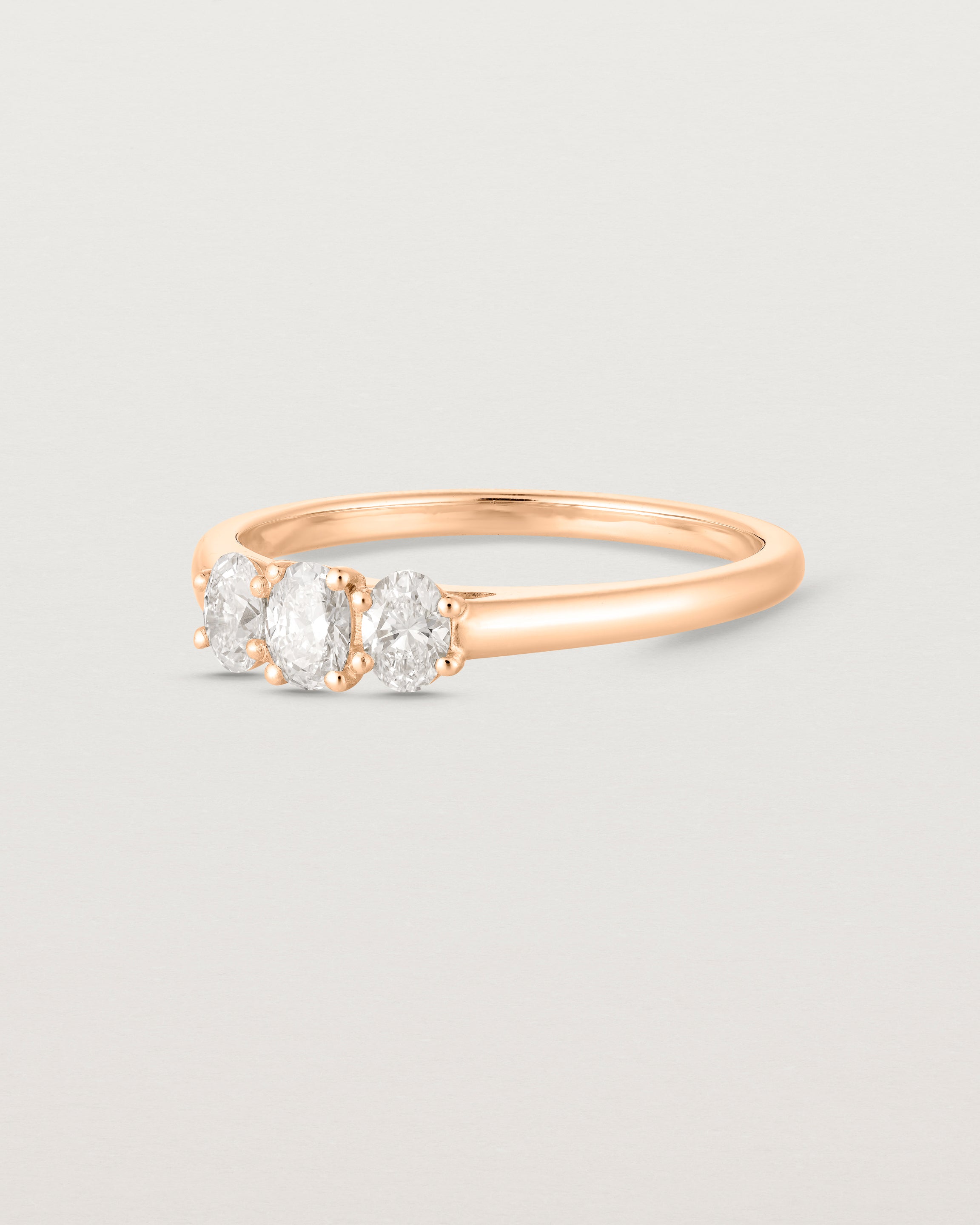 Side on deep etched image of a rose gold engagement ring with three oval white diamonds.