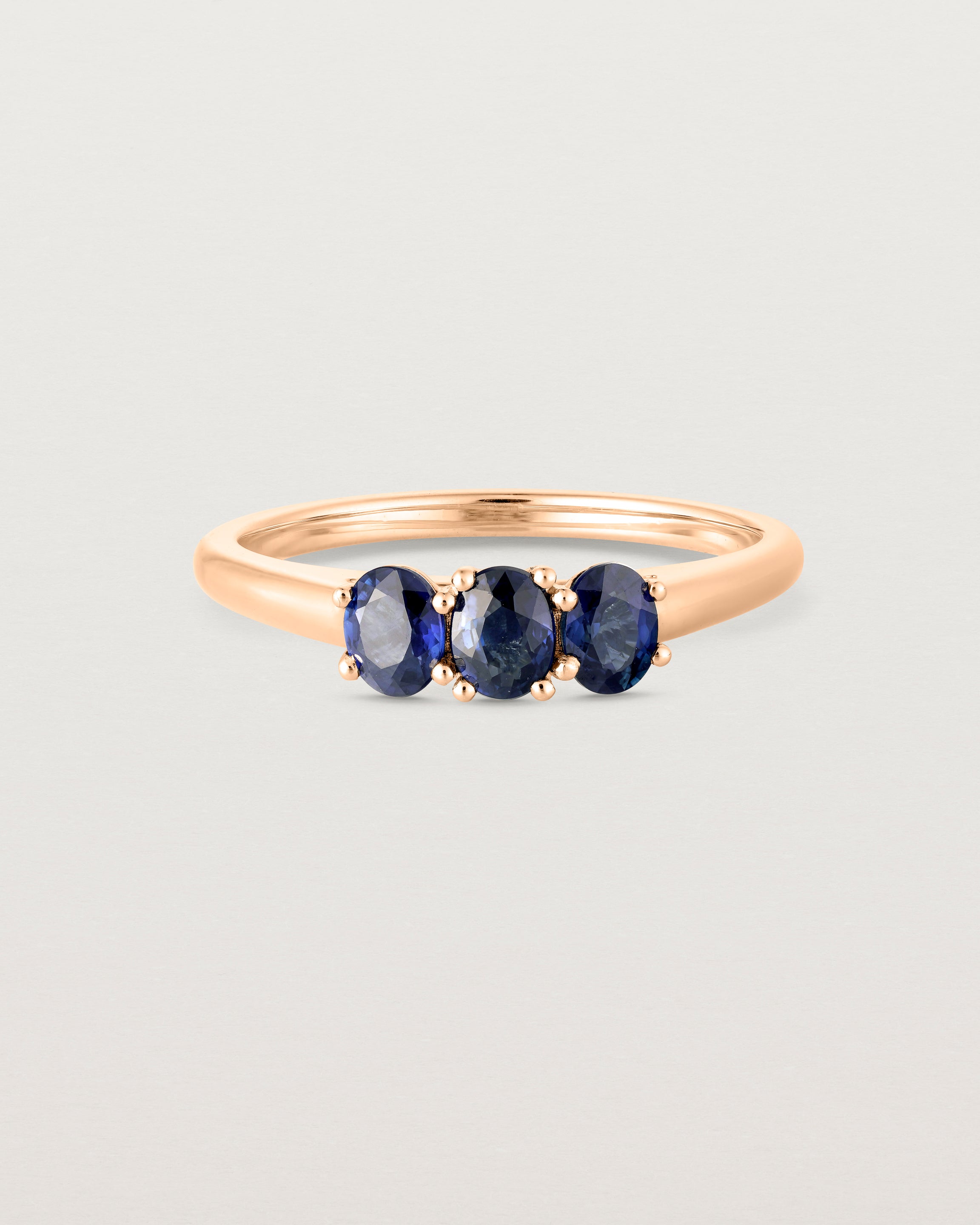 Front on deep etched image of a rose gold engagement ring with three oval sapphire stones.