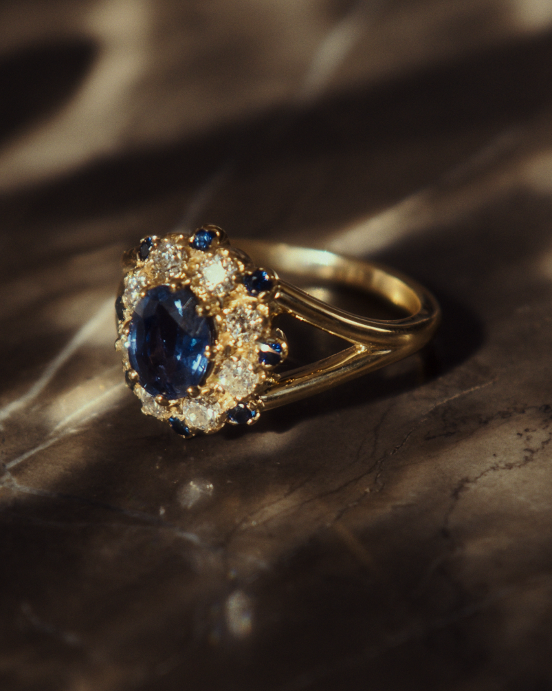 Close up image of a side facing yellow gold vintage inspired ring with an oval Ceylon Sapphire and a tiered halo of Old Cut Diamonds and Australian Sapphires on dark surface.