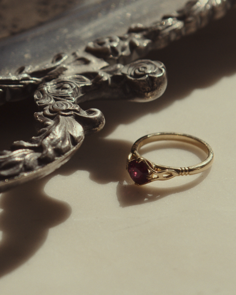 Close up image of yellow gold vintage inspired ring with a cushion cut ruby, flat on table beside silverware.