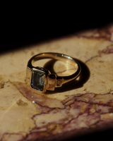 Close up side facing image of yellow gold vintage inspired ring with an emerald cut African parti sapphire with graduating layers either side of the central stone and two white diamond baguettes, on a flat marble surface.