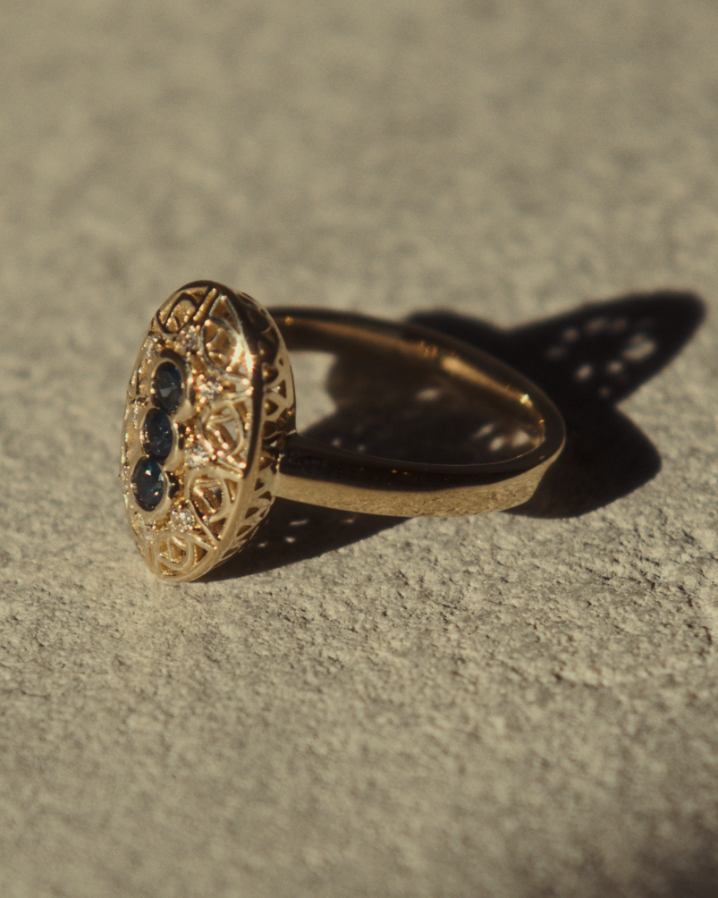 Close up image of a side facing yellow gold vintage inspired shield ring with three Australian sapphires and surrounded by eight old cut white diamonds on flat textured surface.