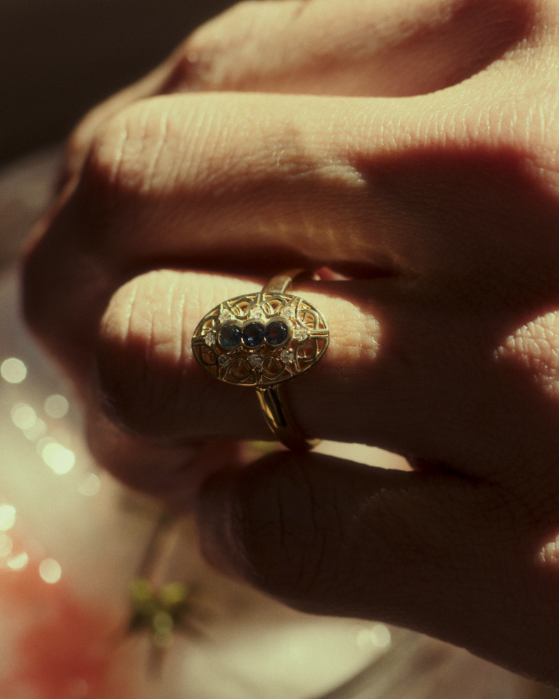 Close up image of a woman wearing a yellow gold vintage inspired shield ring with three Australian sapphires and surrounded by eight old cut white diamonds.