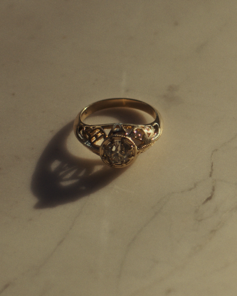 Close up image of a front facing yellow gold vintage inspired ring with old mine cut diamond, framed by an intricate filagree inspired gallery, on flat marble surface.