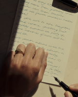 Close up image of a woman wearing a yellow gold vintage inspired ring with old mine cut diamond, framed by an intricate filagree inspired gallery, while writing using calligraphy pen.