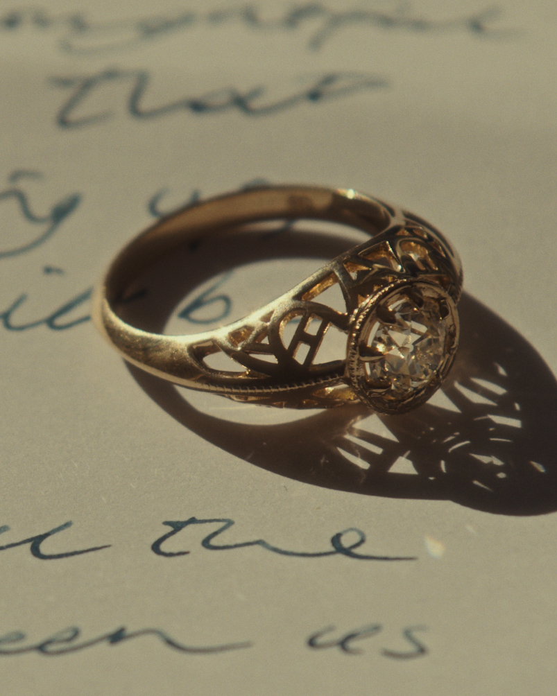 Close up image of a side facing yellow gold vintage inspired ring with old mine cut diamond, framed by an intricate filagree inspired gallery, on paper with text.