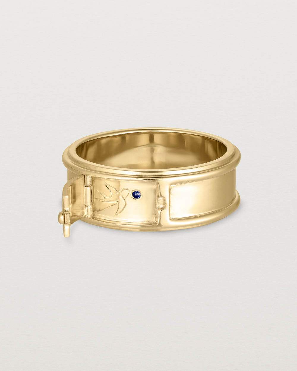 Side facing deep etched image of a yellow gold ring that features a hidden hinge detail, revealing a swallow in flight with a Ceylon sapphire.