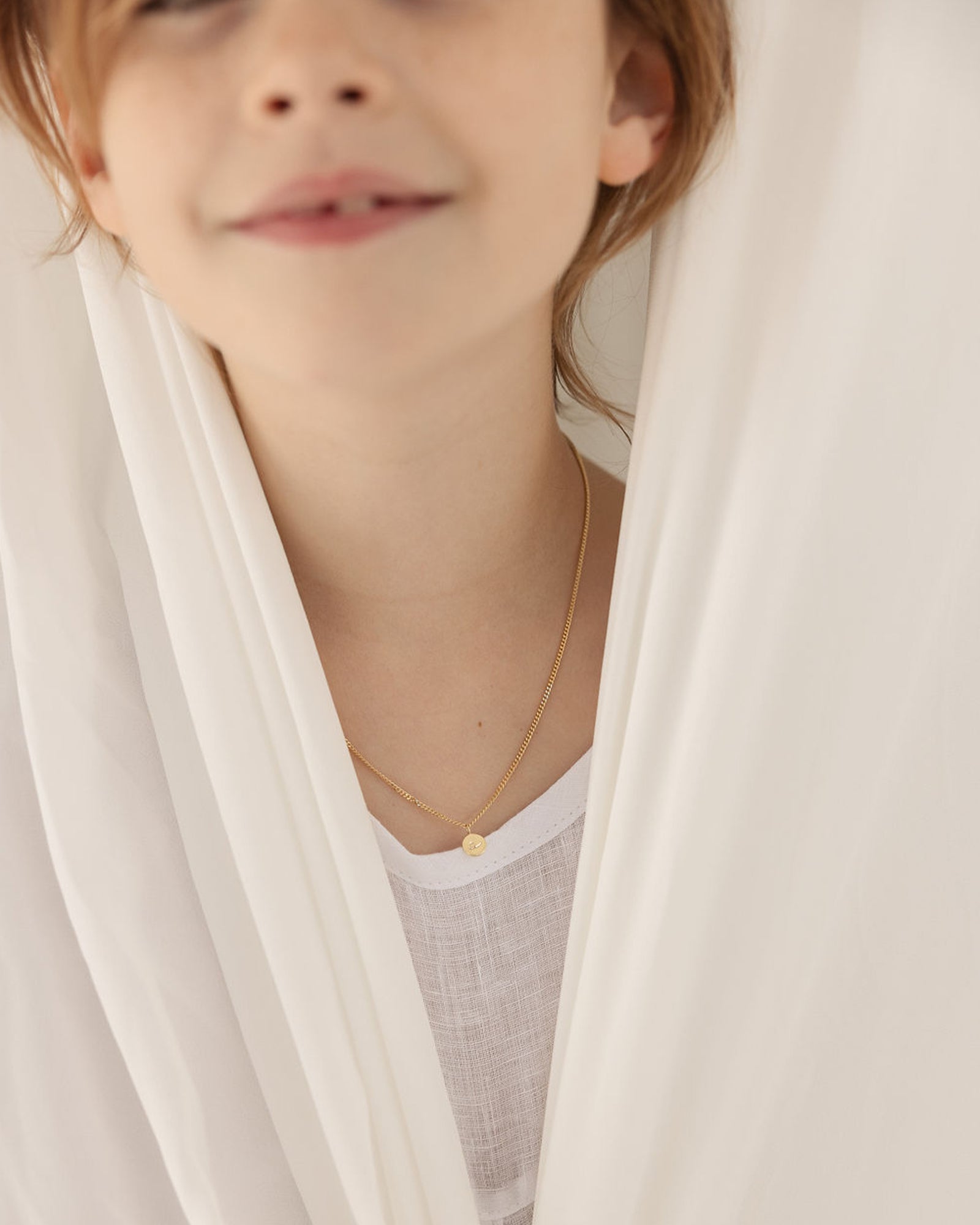 Front on image of the Petit Mae Pendant in yellow gold, shown on a child model peeking behind a sheet