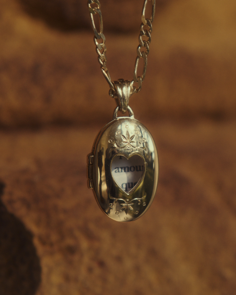 Close up image of a yellow gold chain with locket featuring intricate engraving framing a heart shaped window into the locket.