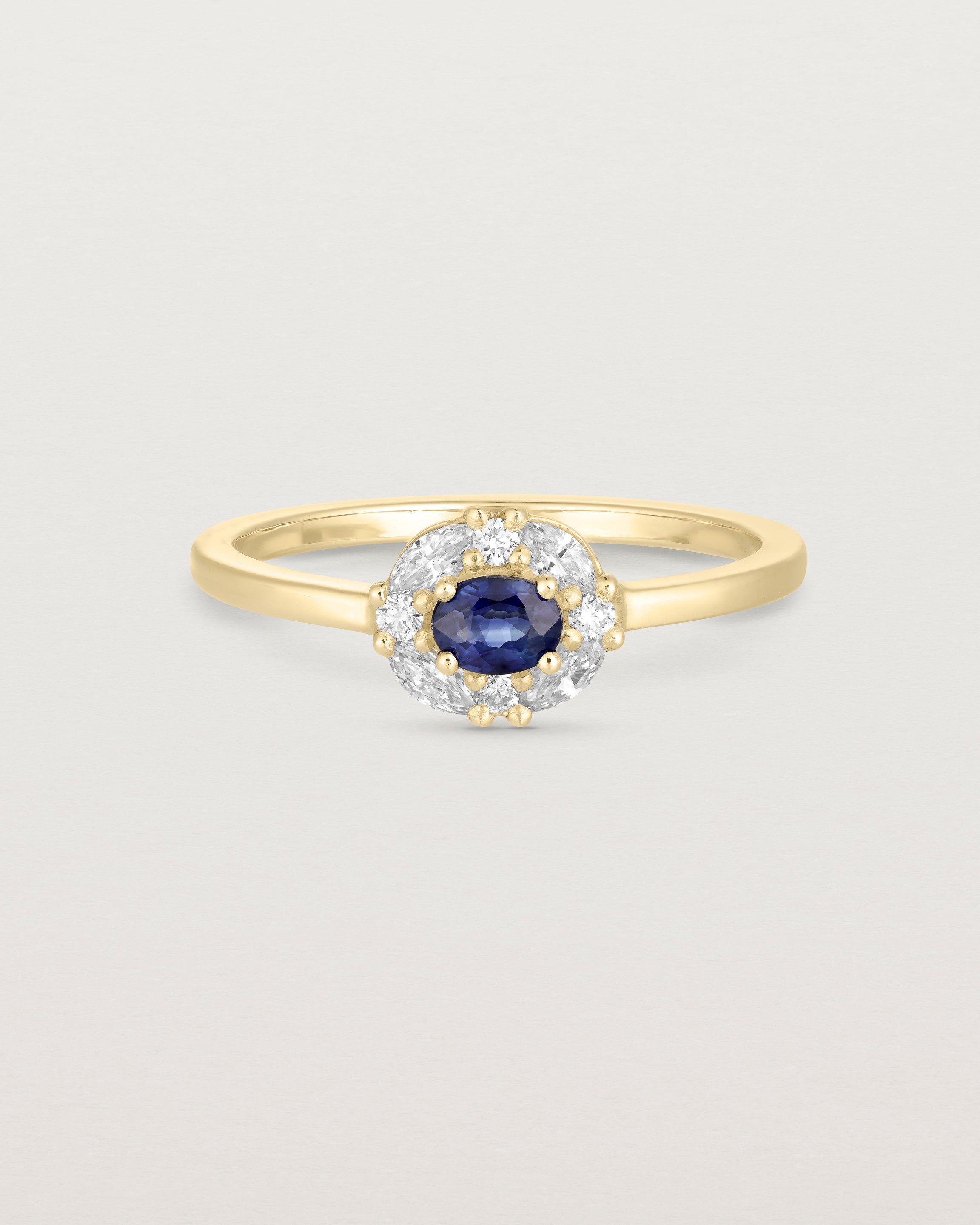 Front on deep etched image of a yellow gold engagement ring with an oval sapphire stone and a halo of eight diamonds.