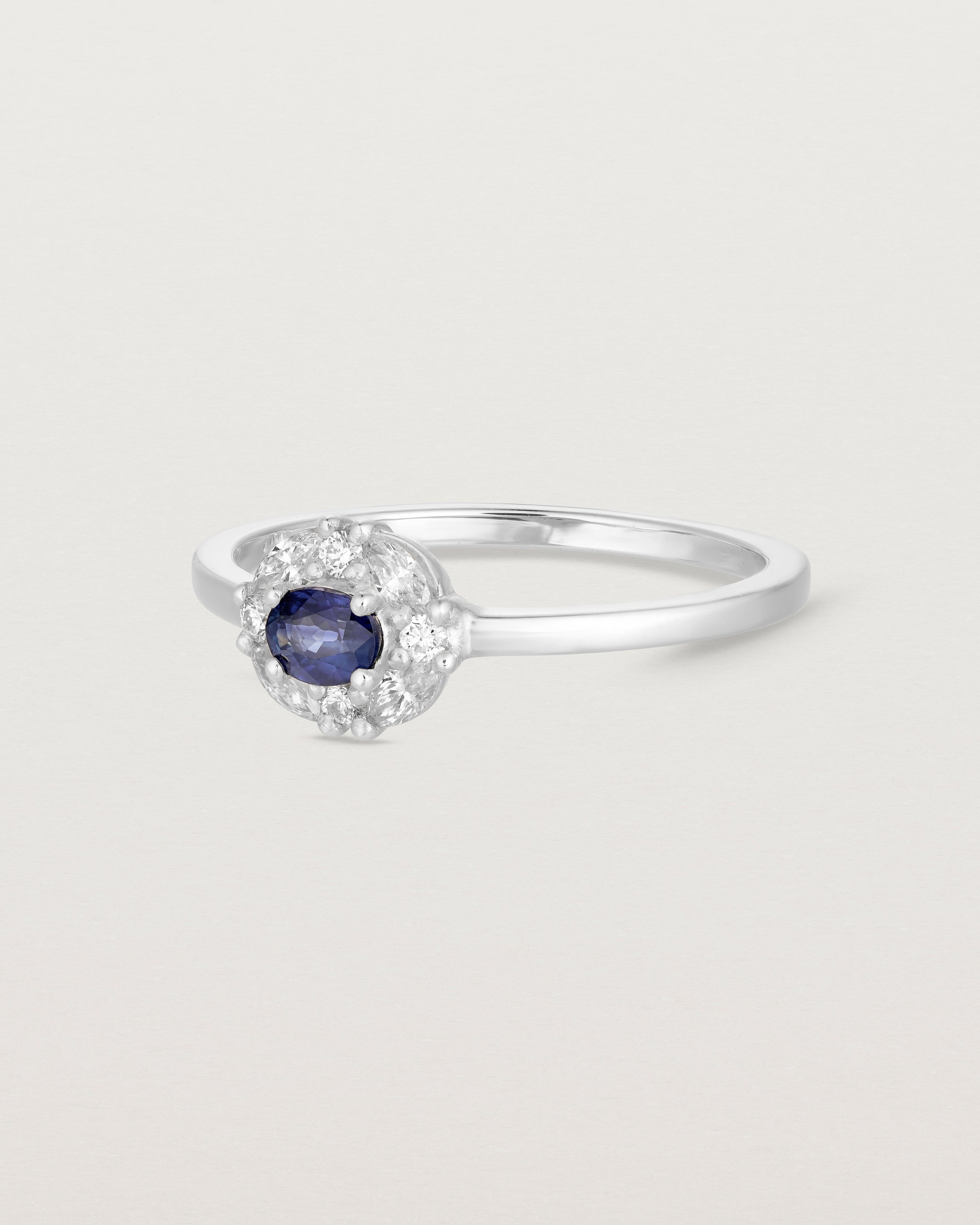 Side on deep etched image of a white engagement ring with an oval sapphire stone and a halo of eight diamonds.