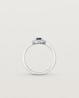 Standing deep etched image of a white engagement ring with an oval sapphire stone and a halo of eight diamonds.